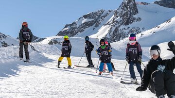 A group of children in ski suits stands on a snow-covered slope. In the background, snow-covered mountains and a clear blue sky can be seen.