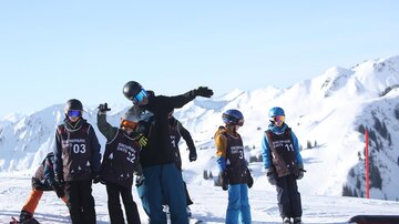 A group of skiers and snowboarders stands on a snow-covered mountain. In the background, majestic mountains and a blue sky can be seen.