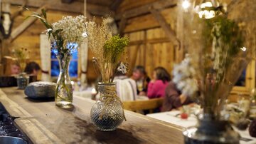 A cozy wooden parlor with a long table and ornate vases filled with dried flowers. In the background, guests can be seen chatting.