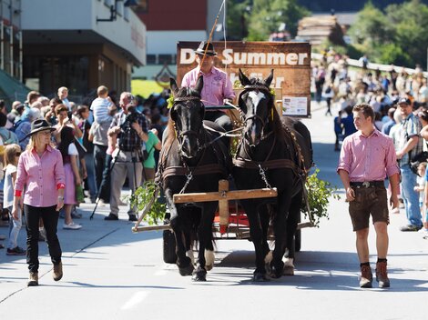 A horse-drawn wagon moves through a busy street. Many spectators in traditional clothing stand along the way and watch the parade.