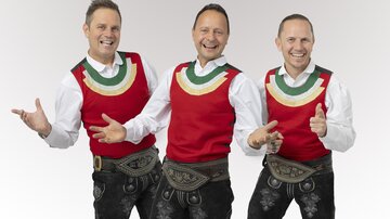 Three men in traditional attire are happily standing together. They are wearing red vests and black leather trousers and exude good cheer.