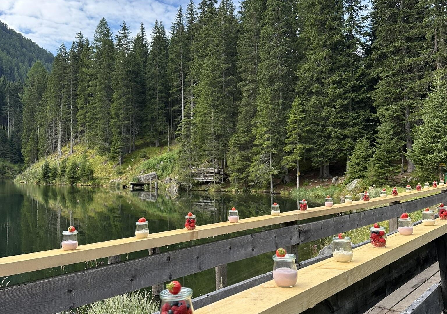 A picturesque wooden boardwalk at the shore of a clear body of water. On the boardwalk, there are glasses with colorful fruits and desserts, surrounded by tall fir trees.