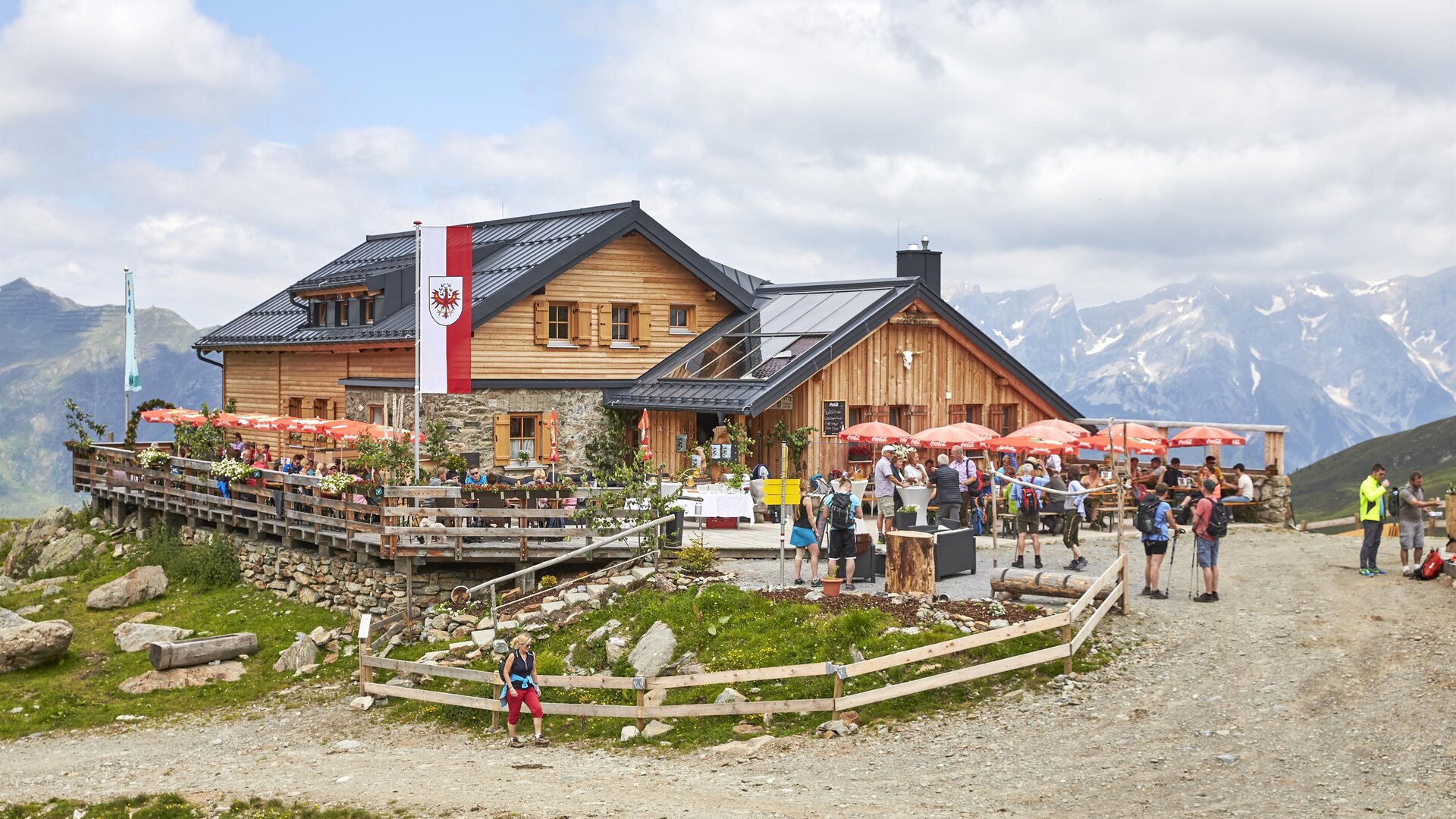 A cozy mountain cabin with a terrace and many visitors. In the background, high mountains and a partly cloudy sky can be seen.