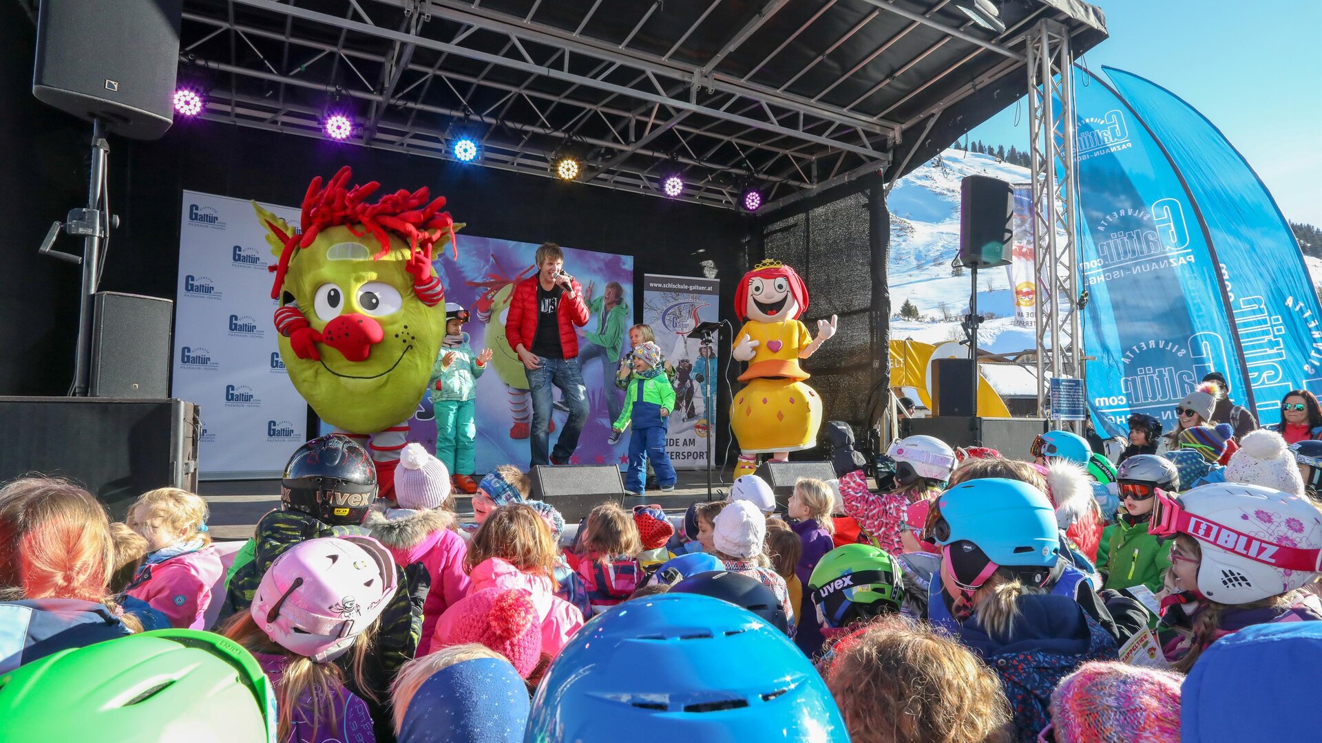 Eine fröhliche Veranstaltung mit Maskottchen auf einer Bühne. Viele Kinder mit Helmen schauen gespannt zu.