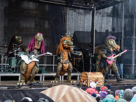 Eine Band von Drachen-Maskottchen spielt auf einer Bühne. Im Hintergrund sind Musikinstrumente und eine begeisterte Menge zu sehen.