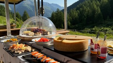 Ein liebevoll angerichtetes Buffet im Freien mit verschiedenen Speisen, darunter Käse, Obst und belegte Brote. Im Hintergrund ist eine malerische Berglandschaft mit Bäumen zu sehen.