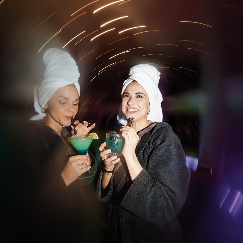 Two women in bathrobes and with towels on their heads are holding colorful cocktails. In the background, lights and a relaxed atmosphere can be seen.