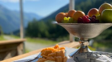 A rustic table with various types of cheese and fresh fruit. In the background, mountains and a clear blue sky can be seen.