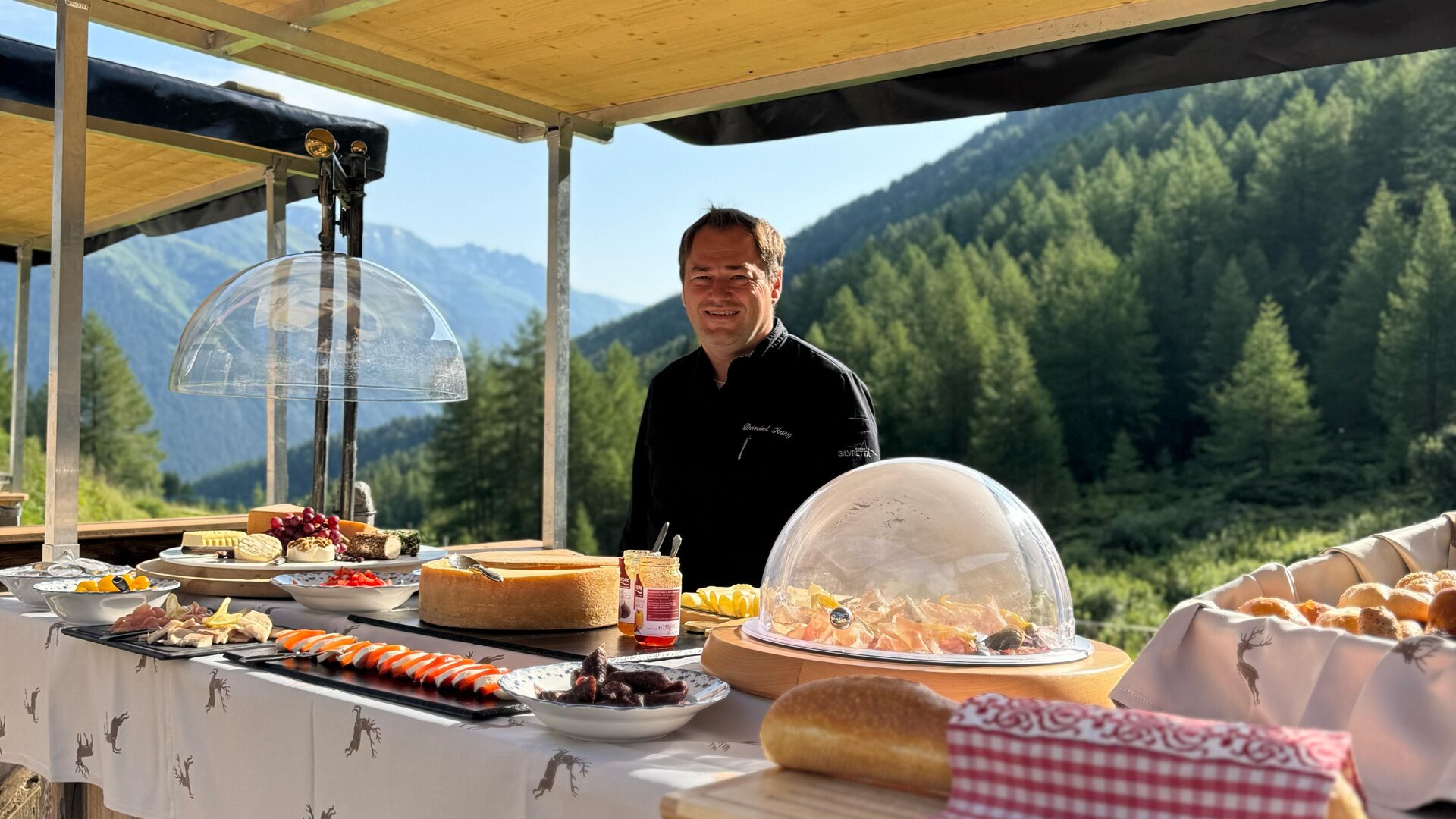 A friendly vendor stands at a wooden stand with a variety of foods. In the background, mountains and forests can be seen.