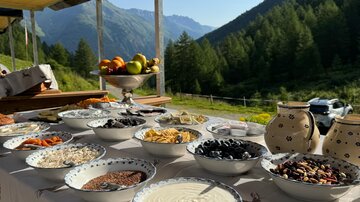 A beautiful table outdoors with various bowls filled with ingredients like yogurt, fruits, and granola. In the background, green mountains and a clear blue sky are visible.