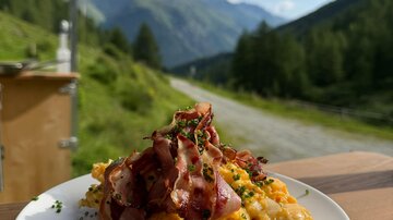 A plate of scrambled eggs and crispy bacon, garnished with chives. In the background, green mountains stretch out under a blue sky.