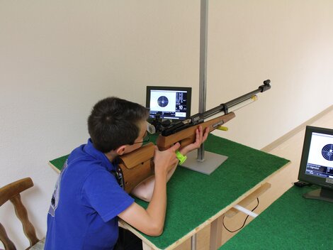 Ein Junge schießt mit einem Luftgewehr auf ein Ziel. Vor ihm sind zwei Monitore, die seine Treffer anzeigen.