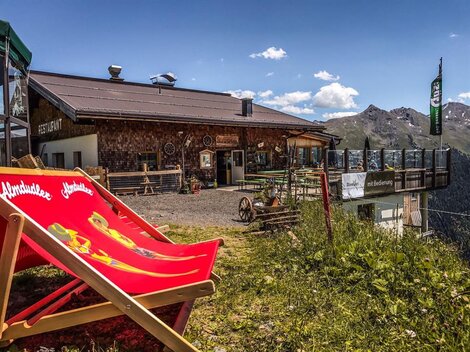 A rustic mountain restaurant with a sun terrace. In the foreground is a red deck chair on a green meadow.