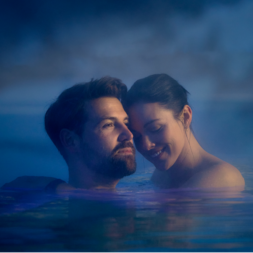 A couple stands in calm water, surrounded by steam. They smile at each other and appear relaxed.