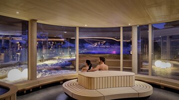 Ein modernes Spa mit großen Fenstern und einer entspannenden Atmosphäre. Ein Paar sitzt zusammen und genießt die Aussicht auf einen beleuchteten Außenbereich.