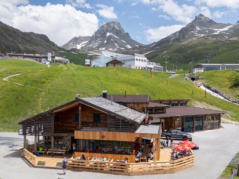 Eine rustikale Hütte in den Bergen mit Terrassenbereich. Im Hintergrund sind grüne Wiesen und schneebedeckte Gipfel zu sehen.