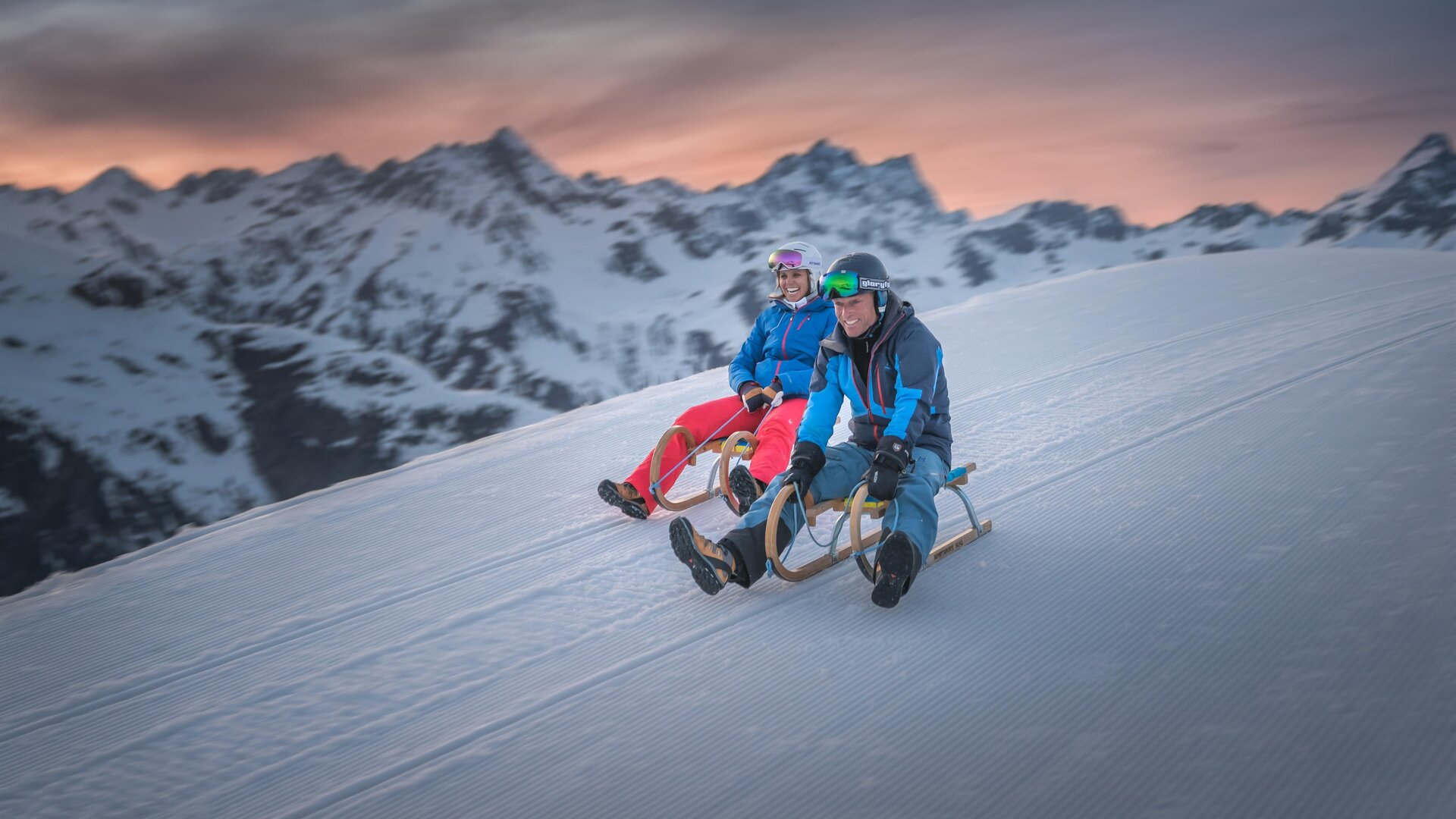 Zwei Personen sitzen auf Schlitten und genießen den Schnee auf einem Berg. Im Hintergrund sind schneebedeckte Berge und ein schöner Sonnenuntergang zu sehen.
