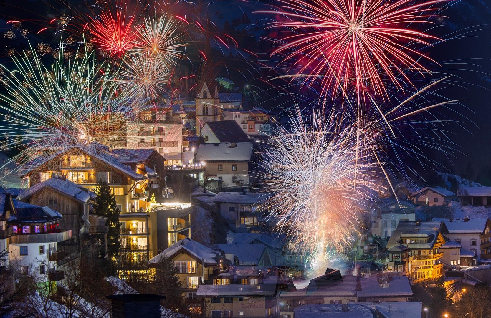 A festive scene with colorful fireworks over a snow-covered village. The houses shine in the light of the fireworks, creating a festive atmosphere.