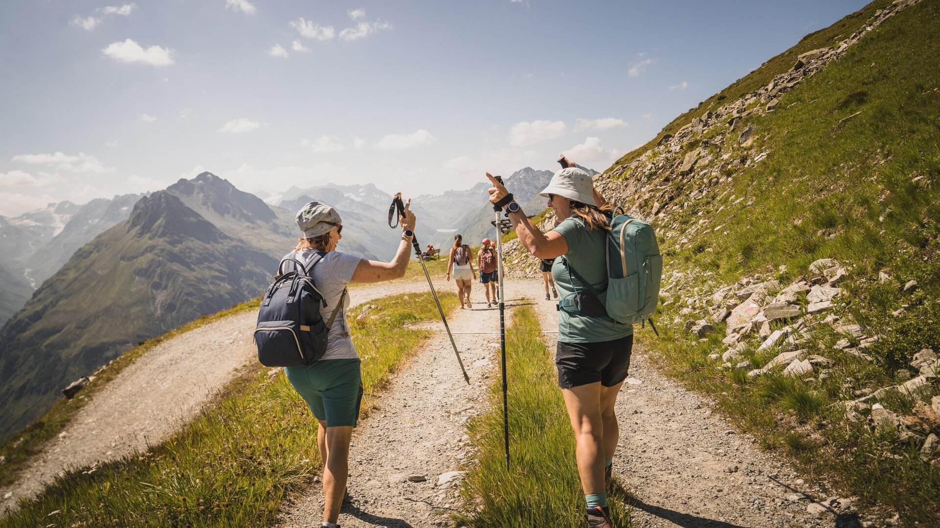 Two hikers on a mountain path enjoy nature. In the background, other hikers and mountains can be seen.