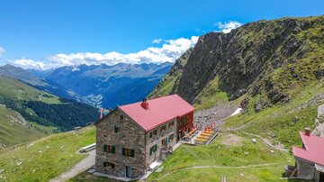 Eine Berghütte mit roten Dächern in einer üppigen grünen Landschaft. Im Hintergrund sind hohe Berge und ein klarer blauer Himmel zu sehen.
