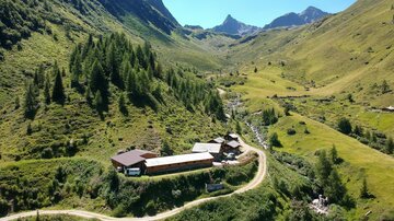 A picturesque mountain landscape with a tranquil valley and green meadows. A small farmhouse lies along a winding road, surrounded by majestic mountains.