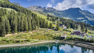 A beautiful mountain landscape with green forests and a clear lake. In the background, majestic mountains and some buildings are visible.