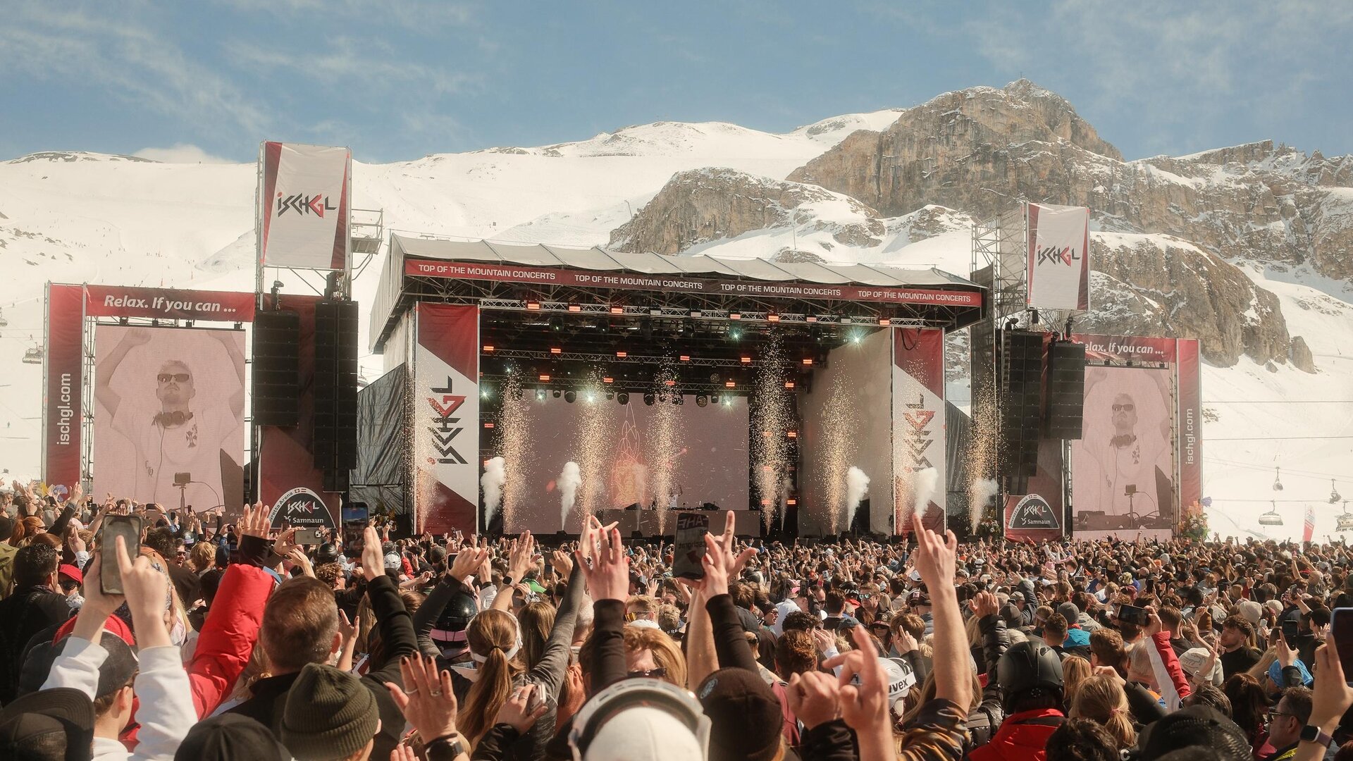 A large music festival in the mountains with many people dancing to the show. In the background, snow-covered mountains and a stage with lights can be seen.