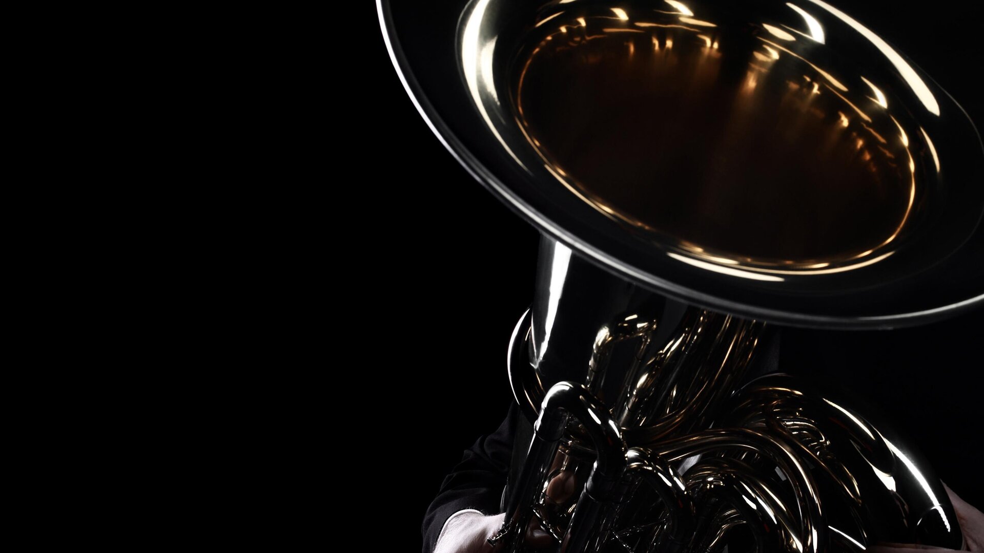 A musician holds a shiny tuba in the dark. Only the lower part of the instrument and the hands are visible.