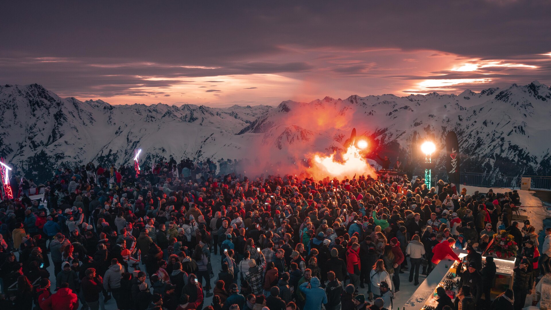 A large crowd gathers in the mountains at sunset. Festive atmosphere with colorful lights and smoke torches.