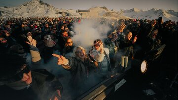 A lively crowd at an event in the mountains. Smoke and snow surround the celebrating people, who are having fun and using their smartphones.