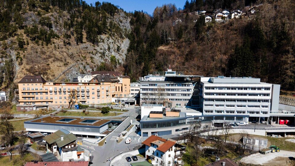 A modern building complex in a mountainous environment. Several buildings are visible, surrounded by trees and hills.