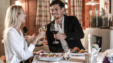 A romantic dinner in a warm, wood-paneled room. A couple toasts with wine glasses, surrounded by an elegant table with delicious food.