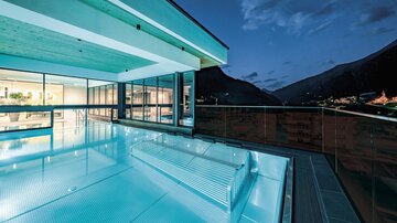 Ein modernes Schwimmbad mit glattem Wasser auf einer Terrasse. Im Hintergrund sind die Berge und der Abendhimmel zu sehen.