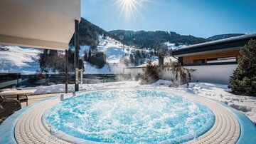 Ein Whirlpool im Freien mit sprudelndem Wasser, umgeben von schneebedeckten Bergen. Die Sonne strahlt am blauen Himmel.
