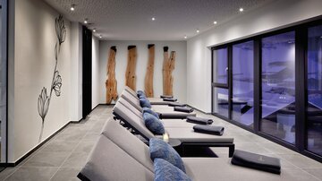 Ein moderner Ruheraum mit Relaxliegen und großen Fenstern. Die Wände sind mit eleganten Holzdetails und einer Blumendekoration gestaltet.