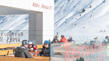 Eine Gruppe von Menschen sitzt in einem Restaurant im Freien, umgeben von schneebedeckten Bergen. Die Sonne scheint und es herrscht eine entspannte Atmosphäre.