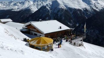 Ein Berghaus in schneebedeckter Landschaft mit Blick auf die umliegenden Berge. Die Terrasse ist mit einem gelben Sonnenschirm ausgestattet und lädt zum Entspannen ein.