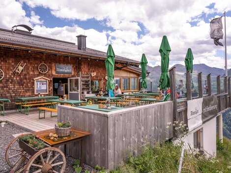 Eine Berghütte mit einem gemütlichen Außenbereich. Es gibt grüne Sonnenschirme und eine schöne Aussicht auf die Berge.