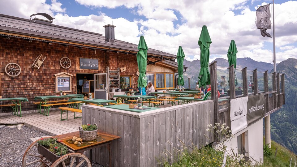 Eine Berghütte mit einem gemütlichen Außenbereich. Es gibt grüne Sonnenschirme und eine schöne Aussicht auf die Berge.