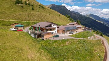 Eine gemütliche Berghütte umgeben von grünen Wiesen und Bergen. Der Himmel ist klar mit wenigen Wolken.