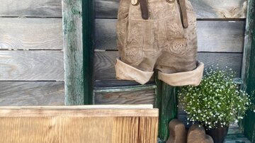 Eine süße, hölzerne Wand mit einem Paar hängender kurzer Lederhosen und flauschigen Blumen. Darunter befindet sich ein Holzschild mit dem Schriftzug „Almstubn“.
