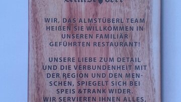 Ein Holzbrett mit einer Begrüßungsbotschaft für ein Restaurant. Der Text spricht von der Verbindung zur Familie und der Bedeutung von Gastfreundschaft.