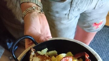 Ein herzhaftes Gericht in einer kleinen Pfanne, bestehend aus Kartoffeln, Speck und einem Spiegelei. Es wird von einer Person serviert.