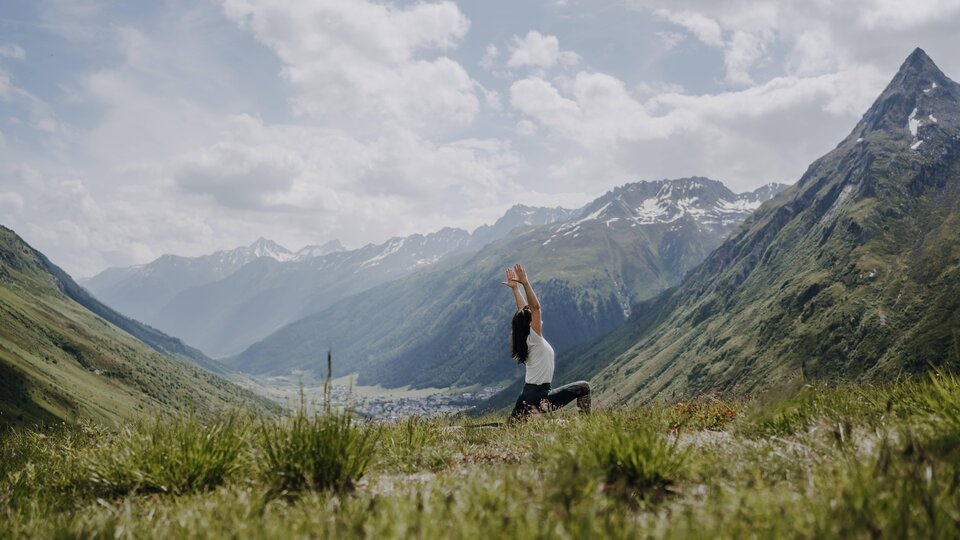 A person is practicing yoga in a picturesque mountain landscape. The surroundings are green and the mountains are partially snow-covered under a clear sky.