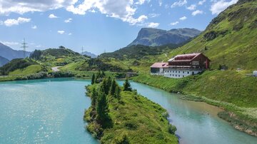Eine malerische Landschaft mit einem klaren, türkisfarbenen See und umliegenden Bergen. Im Hintergrund steht ein traditionelles Gebäude inmitten von grünen Wiesen. | © TVB Paznaun - Ischgl