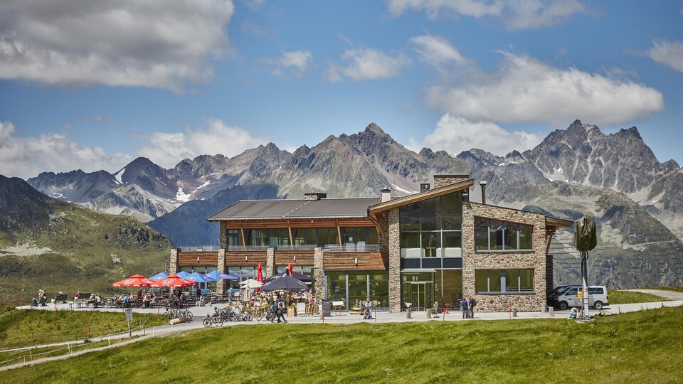 Ein modernes Gebäude in einer malerischen Berglandschaft. Die Terrasse ist mit Gästen und bunten Sonnenschirmen geschmückt.