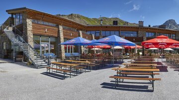 Ein modernes Alpengasthaus mit einer Terrasse und mehreren Tischen. Im Hintergrund sind Berge und ein klarer Himmel zu sehen.