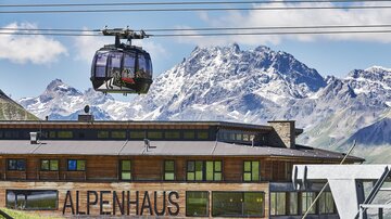 Eine Seilbahn fährt über das Alpenhaus vor beeindruckenden Berggipfeln. Die Landschaft zeigt die Schönheit der Alpen mit klaren Himmel und schneebedeckten Bergen.