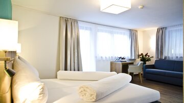A modern hotel room with a large bed, fresh towels, and a cozy sofa. The windows let in plenty of daylight and create an inviting atmosphere.