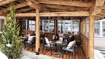 Eine gemütliche Terrasse mit Holzbalken und Tischen. Die Umgebung ist winterlich, mit Schnee und einer grünen Pflanze.